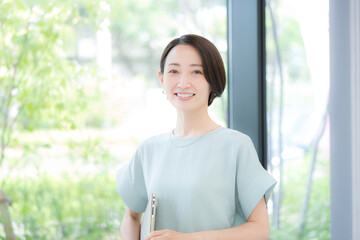 綺麗なオフィスの緑背景でオフィスでパソコンを持つミドルの笑顔のカメラ目線の女性　春夏のオフィスカジュアル