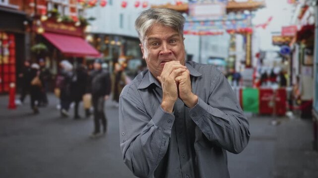 Senior man clasping hands to mouth, gray haired middle age caucasian with worried expression on crowded street with lanterned storefronts; anxiety vulnerability.