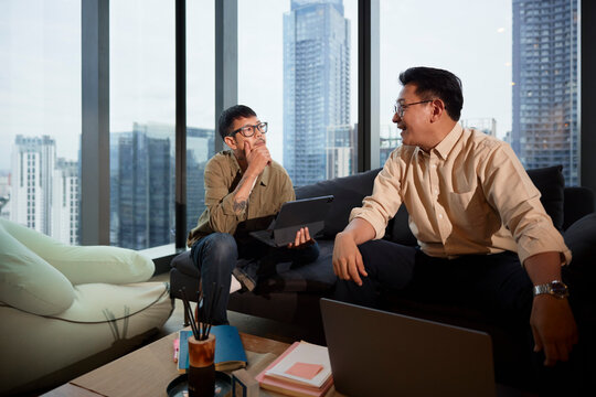 Young male entrepreneur with hand on chin listening to manager while sitting on sofa at office