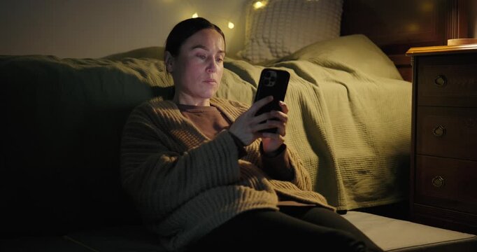 Woman using a smartphone while sitting on the floor in her bedroom at night.