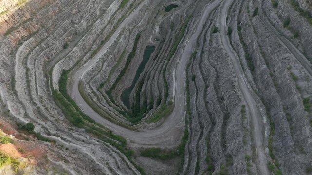 Quarry funnel enclosing elongated water pit with spiraled terraces narrowing inward. Excavation path circling deeply through striped rock layers and green edging. Mining trench forming nested curves