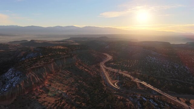 A cinematic drone shot captures a winding road along the rugged mesa ridges of Los Alamos at sunset. Golden hour light flares over the cliffs and Jemez Mountains in this travel transition.