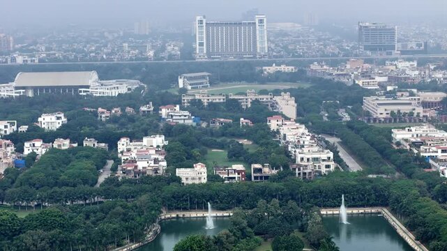 Drone footage of low-rise housing and green pockets framed by elevated roads and distant towers, softened by smog that highlights ongoing air pollution in Greater Noida.