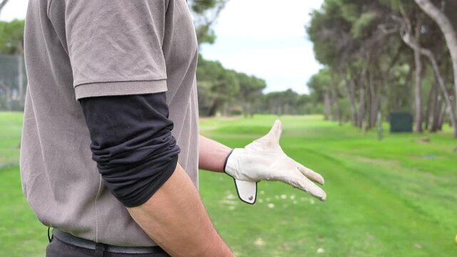 A man in a grey shirt and black sleeves is standing on a golf course. He is wearing gloves and has his arm around his elbow