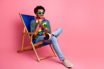 Young man relaxing in a blue beach chair wearing sunglass and a colorful lei on a pink background