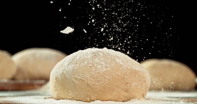 Cinematic moving shot of leavened dough isolated on black background with falling flour in super slow motion, 1000 fps. Dynamic baking concept, artisan bread preparation and texture detail.
