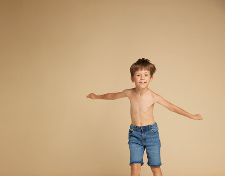 Full length image of a boy at school jumping up with raised hands, dressed only in jeans shorts, and bare feet, isolated beige background. Copy space