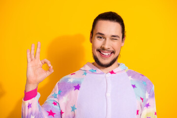 Happy man wearing pastel starry pajama winking and giving ok sign against yellow background