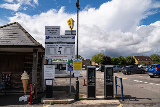 Bourton-on-the-Water, United Kingdom, 12 July 2023, A car park features pay stations and visible parking signs. Vehicles occupy marked spaces under bright summer clouds.