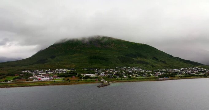 Sortland town sits on calm coastal waters beneath green mountain wrapped in low clouds and mist, aerial orbit establish