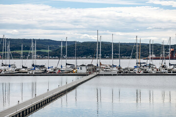 La Baie, Quebec, Canada &ndash; August 19, 2025: Wooden pier leading to a marina with moored sailboats on calm water under a cloudy summer sky, with distant hills and shoreline visible in the background