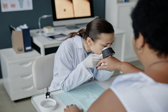 Young adult Asian woman dermatologist examining hand of middle aged Black woman patient using dermatoscope during dermatology consultation in medical office
