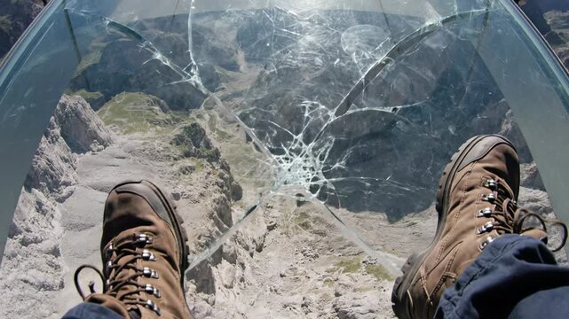 A conceptual POV representation of extreme heights and modern anxiety showing hiking boots on a cracked glass observation deck looking down into a deep mountain valley. Vertigo.