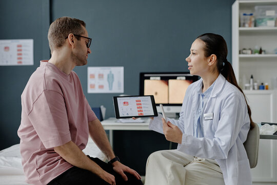Caucasian young adult man consulting with Asian young adult female dermatologist holding digital tablet displaying skin analysis results in medical office during dermatology appointment