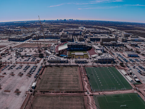 Aerial view of a vibrant stadium surrounded by sports fields and parking lots, juxtaposed against the distant city skyline, Frisco, Texas, United States.