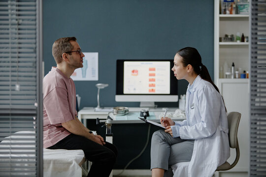 Caucasian middle aged man sitting on examination table consulting with Asian young adult woman dermatologist holding clipboard, in medical office with computer monitor in background