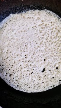 Vertical, macro top-down view of yeast pancake batter cooking in a hot black pan. The mixture sizzles and forms a porous, bubbly texture as it fries, showing the complete culinary process.