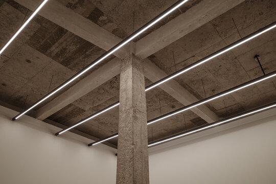 A modern interior shot featuring a raw concrete pillar and ceiling with exposed structural beams. Bright linear LED light fixtures are suspended in a parallel arrangement.