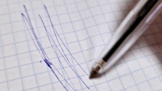 Macro close-up shot of a ballpoint pen tip drawing random zigzag lines with blue ink on a sheet of white squared paper. The detail highlights the texture of the paper and the ink being applied.