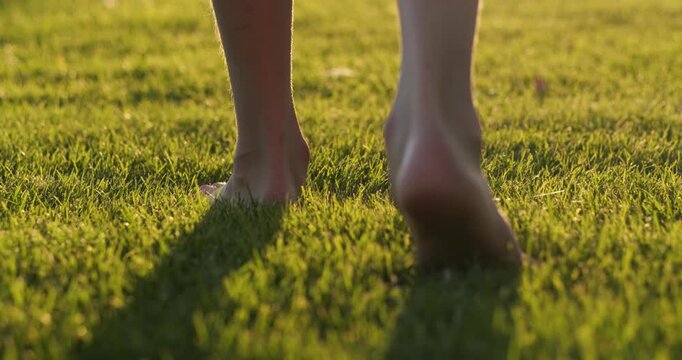 Barefoot person walks on fresh grass during golden hour at sunset. Barefoot steps touch nature softly and natural. Sunlight and warm atmosphere. Wellness, slow living and outdoor relaxation themes.