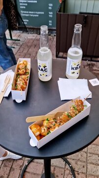 High-angle shot of skewered dumplings in paper trays topped with green onions and nuts beside two bottles of lemon Kvas. The meal sits on a black round table at outdoor urban cafe near a menu board.