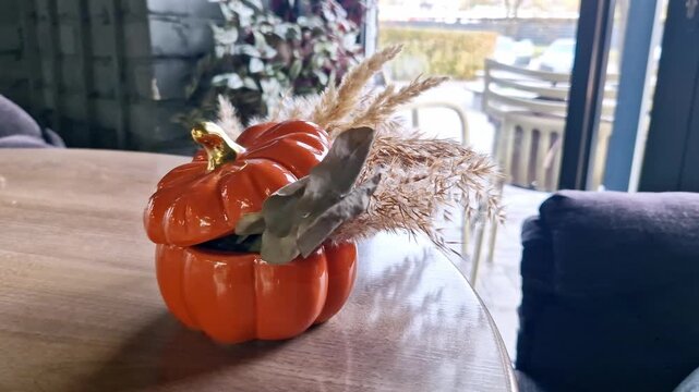 Close-up shot of an orange ceramic pumpkin with a gold stem acting as a vase for dried pampas grass. The autumn centerpiece sits on a round table near a large window.