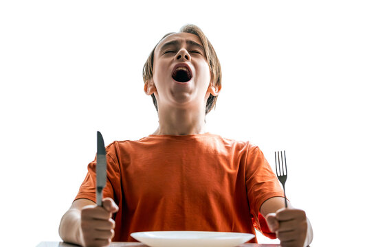 Angry hungry boy child waiting for dinner. Holding fork and knife in his hands. Little boy sits in front of an empty plate. Upset little boy waiting for dinner while holding a fork, spoon. Hungry boy