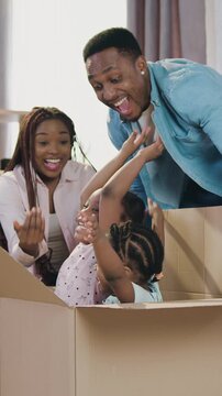 Good-looking smiling modern young dark-skinned man and woman playing with their kids which jumping out from carton box after they moved into newly acquired flat and unpacking boxes,slow motion