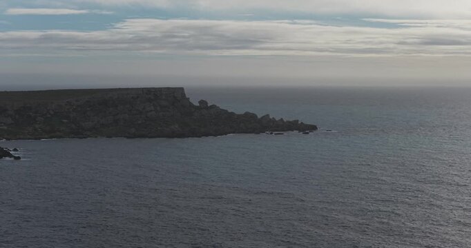Aerial view of the rugged Ras in-Niexfa coastline with dark rocks contrasting against the shimmering sea under a cloudy sky, Ras in-Niexfa, Mellieha, Malta.