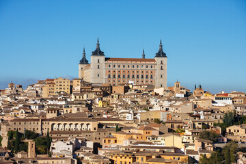 The Alcázar of Toledo, a historic fortress and palace, stands prominently atop a hill overlooking the densely packed cityscape of Toledo, Spain, under a clear blue sky. © Nick