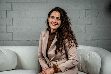 Professional woman smiling, posing for a portrait in a modern office space, sitting on a sofa with a concrete wall background