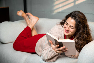 Happy woman lying on a comfortable sofa reading a book, embracing quiet relaxation and a cozy lifestyle
