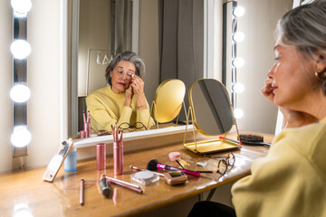 Senior woman sitting at her vanity, carefully removing eye makeup with a cotton pad, reflecting in the mirror