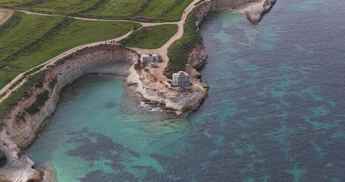 Aerial view of the rugged coastline meeting the clear turquoise sea, contrasting with the green fields above, Munxar Path, Marsaskala, Malta.