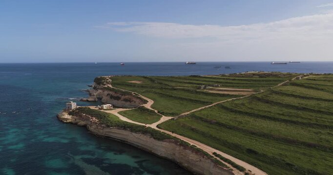 Aerial view of buildings on the rocky coast, the turquoise sea contrasting sharply with the green fields, Munxar Path, Marsaskala, Malta.