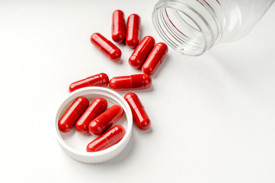 Red capsules spilled from an open plastic container on a white background, photographed as a clean medical still life.

