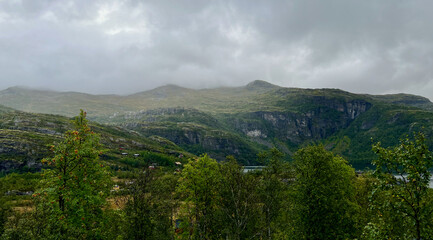 Dramatic mountain landscape in Myrdal, Norway featuring steep rocky cliffs, lush green valley, scattered cabins, and misty clouds rolling over the peaks, showcasing the raw beauty of Scandinavian natu © Noppadol