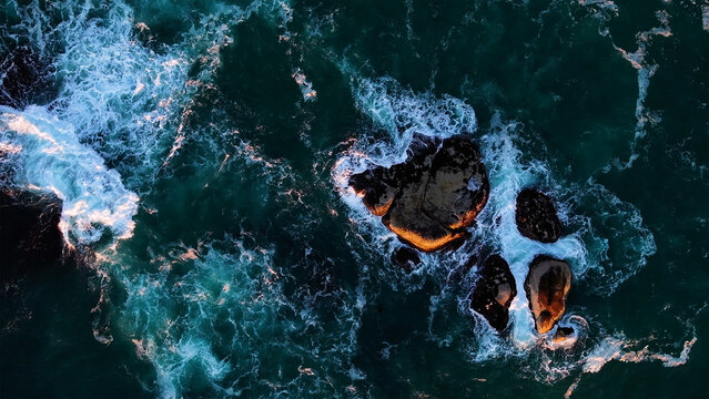 Aerial view of the rugged coastline where the turbulent turquoise ocean clashes against the dark, sun-kissed rocks, Cape Town, Western Cape, South Africa.