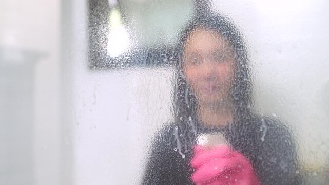 Asian woman in pink gloves using a squeegee to clean soapy shower glass. Bathroom housekeeping concept.