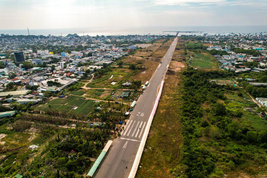 Aerial view of a runway stretching towards the sea, flanked by verdant foliage and urban development, a blend of natural and built environments, Duong Dong, Phu Quoc, Vietnam.