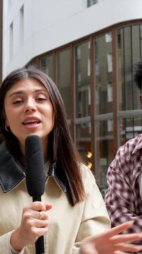 Charismatic female reporter holding a microphone while conducting a street interview with a smiling man, both looking at the camera and engaging with the audience in an urban city setting