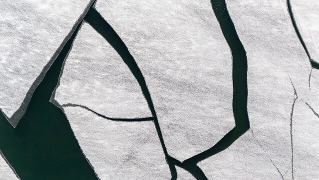 Aerial view of fractured ice floes drift on the dark waters of Lake Superior, creating a stark contrast between frozen white expanses and deep blue depths. Superior, Wisconsin, USA.