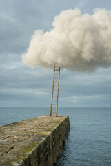 Fototapeta premium A wooden ladder leads from a stone pier into a fluffy white cloud over the ocean.