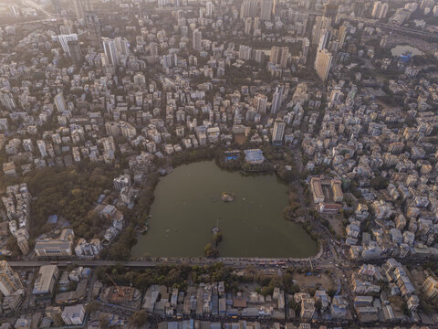 Aerial view of a bustling city surrounding a serene, green-tinged lake, contrasting with dense buildings and roads, captured in the soft, golden light of dawn, Thane, Maharashtra, India.