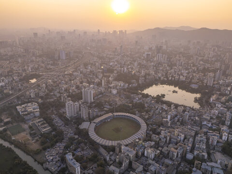 Aerial view of the Dadoji Kondadev Stadium and Talao Pali Lake amidst the urban sprawl, bathed in the hazy glow of the setting sun, Thane, Maharashtra, India.
