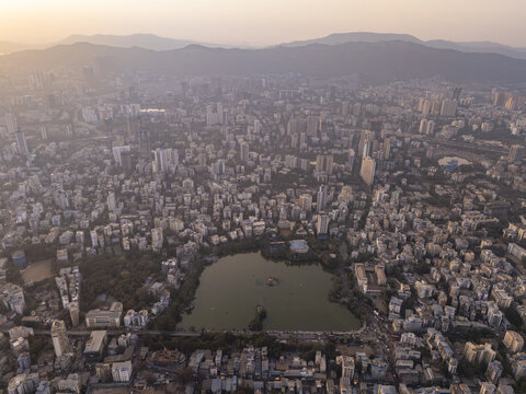 Aerial view of a dense cityscape punctuated by the serene Masunda Lake, with warm sunlight casting a golden hue over the urban sprawl, Thane, Maharashtra, India.