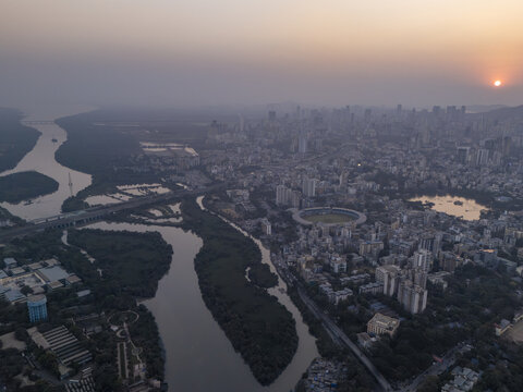Aerial view of the dense cityscape meeting the winding river, with the sun setting over the horizon casting a warm glow, Thane, Maharashtra, India.