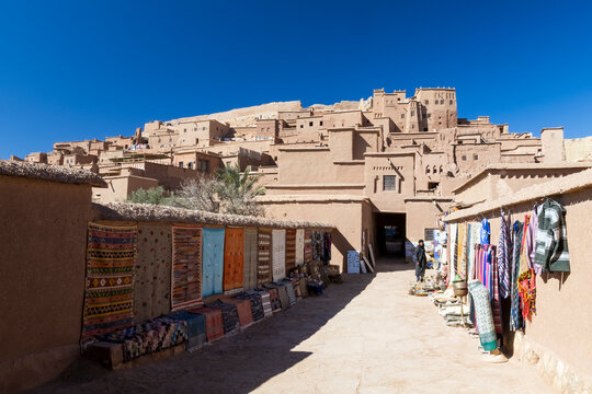 Ait Benhaddou Kasbah with market stalls selling colorful textiles under a clear blue sky. Kasbah ait ben haddou,Marrakech,Morocco