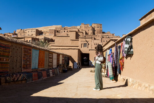 A woman stands in a market street lined with rugs and textiles, with a kasbah in the background. Kasbah ait ben haddou,Marrakech,Morocco