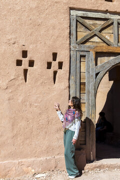 A woman stands by a mud wall with geometric designs and a wooden door frame. Kasbah ait ben haddou,Marrakech,Morocco